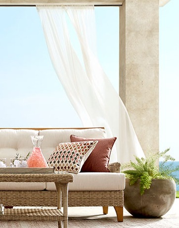 Tan wicker sofa with colorful throw pillows next to white outdoor curtain